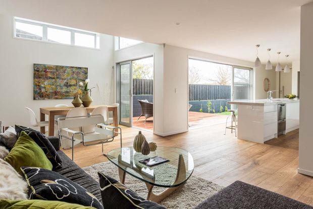 Bright open-plan contemporary living, dining and kitchen area with timber flooring, white walls, extensive glazing and a central white kitchen island. The room opens to a paved outdoor courtyard, while a sectional sofa, glass coffee table, dining table and pendant lights furnish the space.