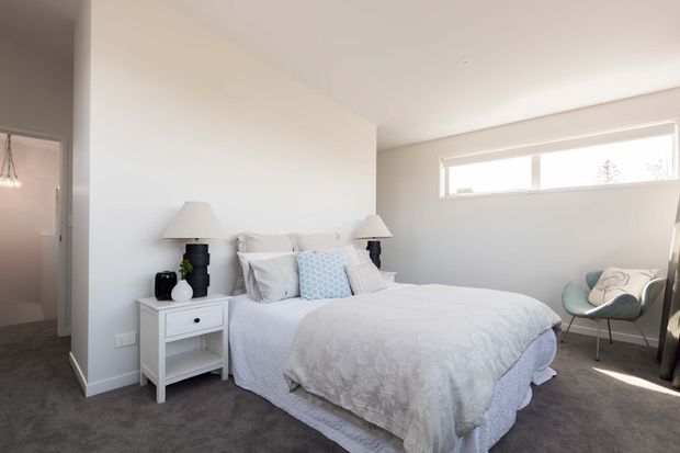 Bright, uncluttered bedroom with a neatly made upholstered bed, white and pale blue bedding, matching bedside tables and lamps, a small accent chair, white walls, a narrow horizontal window, and dark grey carpet. A partially visible adjoining bathroom or hallway is seen through the doorway on the left.