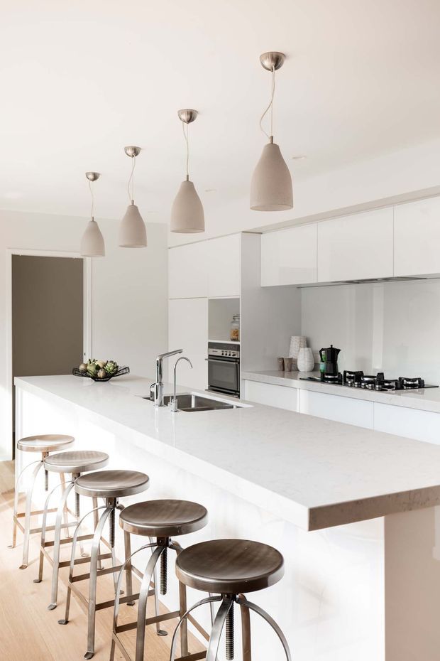 Bright contemporary white kitchen with a large waterfall-edge island, integrated sink and chrome tap, five industrial-style metal bar stools, white cabinetry, pale stone benchtops, and a white tiled or glass splashback. Several neutral pendant lights hang above the island, with built-in appliances and a gas cooktop along the rear wall.