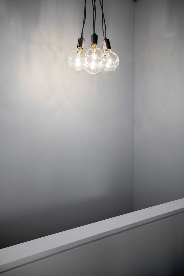 Minimalist interior detail featuring a cluster of three clear spherical pendant lights with exposed bulbs and black cords, suspended against smooth light-grey walls. A white angular ledge or stair balustrade crosses the lower foreground, creating a stark geometric composition with soft reflected light.