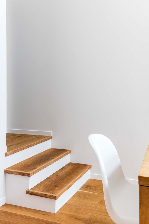 Bright minimalist interior view of a compact stairway with open-riser timber stairs, white painted walls and a white sculptural chair. Timber flooring continues around the staircase, with a partial timber built-in surface visible at the right edge.