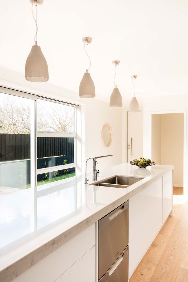 Bright, contemporary white kitchen with a long island featuring an integrated double stainless-steel sink and dishwasher, white cabinetry, and a pale stone benchtop. Four neutral fabric pendant lights hang above the island. Large windows provide abundant natural light, with pale timber flooring and a simple round wall mirror adding warmth.