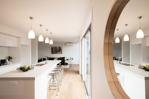 Bright open-plan kitchen and living area with a long white island, integrated white cabinetry, pale timber flooring, and a row of pendant lights. Bar stools line the island, while full-height glazing opens toward the exterior. A large circular timber-framed mirror or opening is visible on the right, reflecting the kitchen and dining area.