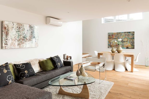 Bright, open-plan contemporary living and dining area with white walls, warm timber flooring, a dark sectional sofa, glass coffee table, sculptural dining chairs, and abstract artwork. High windows provide natural light, creating an airy and inviting interior.