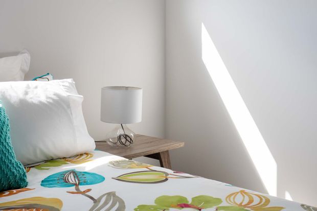 Bright, minimalist bedroom detail with white bedding, a colorful botanical-patterned duvet, a turquoise knitted cushion, and a simple timber bedside table with a white lamp. Strong angular sunlight falls across the plain light wall, creating a calm and airy atmosphere.