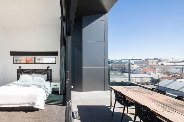 A bright contemporary bedroom opening directly onto a rooftop terrace or balcony. The bedroom has a white bed, dark textured feature headboard, pale walls, and carpeted flooring. Large glazed sliding doors connect the bedroom to an outdoor dining area with a long timber table and black chairs. The terrace has dark exterior cladding and overlooks a dense urban industrial/commercial setting.