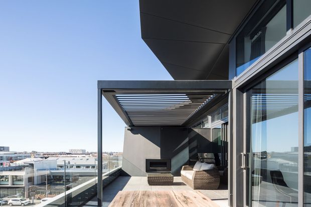 Modern elevated balcony terrace with timber-look decking, glass balustrades, dark architectural cladding, extensive sliding glazing, and a partially covered outdoor lounge area. A slatted pergola provides shade over woven seating and an outdoor fireplace, with an urban view beyond.