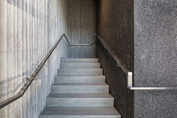 A narrow, straight interior stairway rises between heavily textured concrete walls. The steps are finished in grey concrete, with simple dark metal handrails on both sides and a small landing at the top. The composition is stark, monochromatic, and highly architectural, with no visible furnishings or windows.