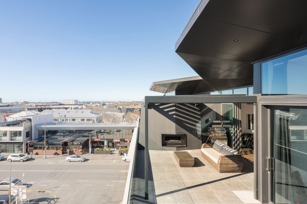 A contemporary rooftop terrace or balcony overlooking an urban commercial streetscape. The outdoor space has large-format light stone or concrete-look paving, a built-in dark fireplace or barbecue feature, woven outdoor seating, glass balustrades, dark metal framing, and a deep cantilevered roof canopy. Strong afternoon sunlight creates patterned shadows across the terrace.