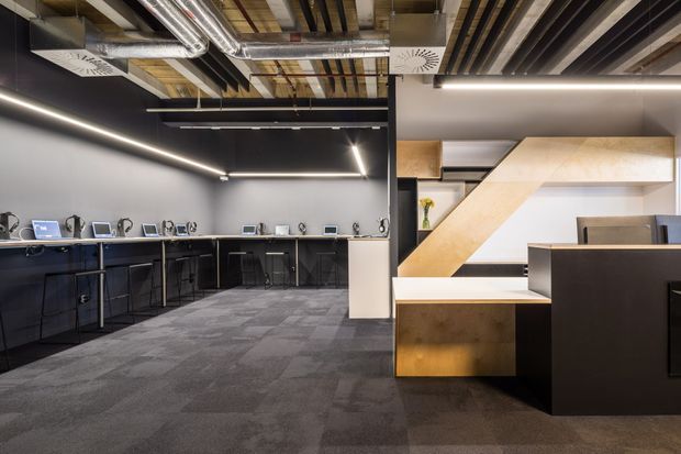 A modern commercial office interior with an open kitchenette or breakout area along the left wall and a large angular timber-clad stair or architectural partition on the right. The space features dark built-in cabinetry, grey carpet tiles, exposed services and timber ceiling elements, linear LED lighting, and a monochromatic industrial fit-out.