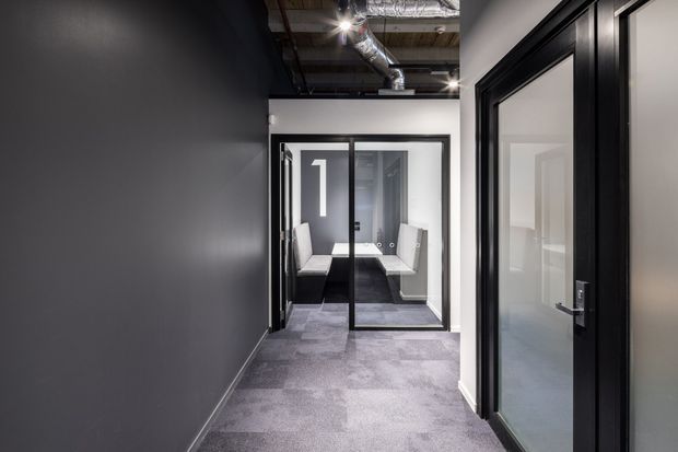 A modern commercial office corridor with dark charcoal walls, black-framed glazed doors, and grey carpet flooring. A small meeting or waiting area is visible through the glass at the end of the hallway. The ceiling is exposed, showing ductwork and services, with recessed and spot lighting.
