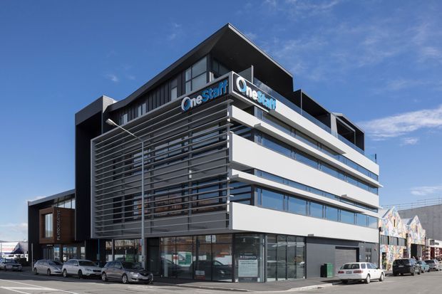 Exterior view of a contemporary multi-storey commercial office building on a street corner. The building features a crisp white and dark charcoal facade, extensive horizontal glazing, prominent external aluminium louvre screens, projecting box forms, and rooftop signage. Cars and neighbouring commercial buildings are visible at street level.