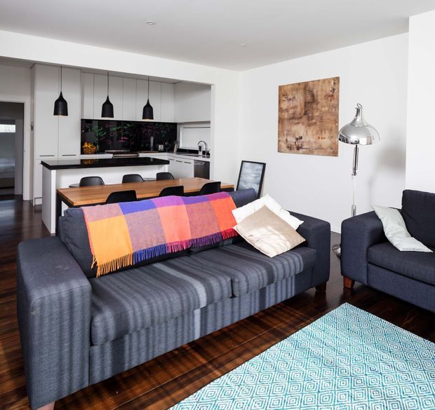 A bright open-plan living and dining area with a contemporary kitchen in the background. The room features charcoal upholstered sofas, a colourful striped throw, patterned rug, dark timber flooring, white walls and cabinetry, black pendant lights, a timber dining table, and a black glossy splashback. A chrome floor lamp and abstract artwork add decorative accents.
