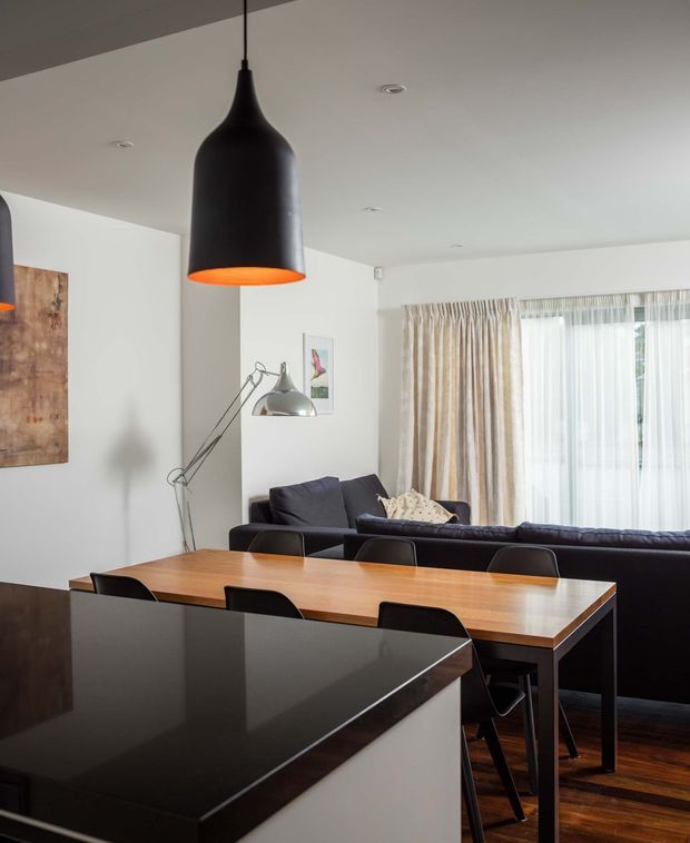 Contemporary open-plan dining and living area viewed from a kitchen island. A long timber dining table with dark dining chairs sits in front of a dark sectional sofa, with a large black pendant light overhead, an adjustable floor lamp, white walls, sheer curtains, and dark timber flooring. A glossy dark benchtop occupies the foreground.