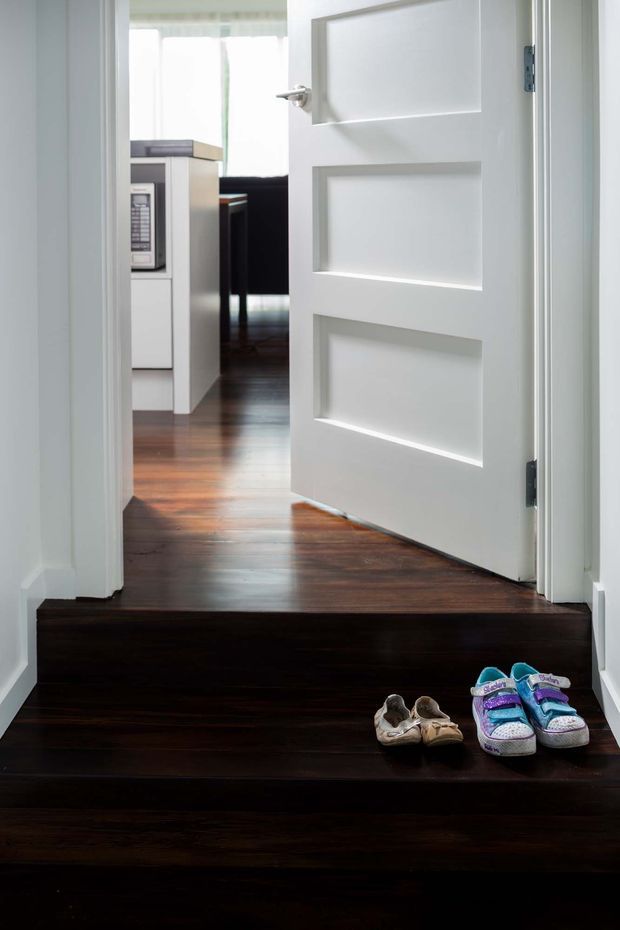 Bright residential interior view looking up dark timber stairs toward an open white paneled door and a kitchen or living area beyond. The stair and upper floor have rich dark wood flooring, while the surrounding walls, trims, and door are white. Children's shoes are placed on the stair, adding a lived-in domestic detail.