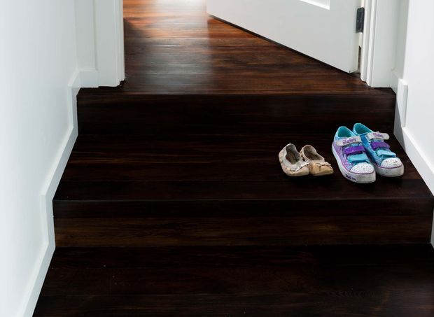 A narrow interior stairway with dark timber stair treads and a matching dark timber landing, enclosed by white walls and white door trim. An open white door leads to the upper landing, with several pairs of shoes placed on the steps.