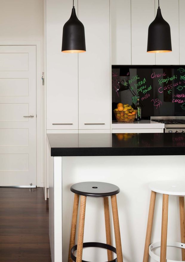 A contemporary kitchen with a black stone island benchtop, white full-height cabinetry, and a glossy black splashback used as a chalkboard. Two oversized matte-black pendant lights hang above the island, which is paired with contrasting black and white timber-legged stools. Dark timber flooring and a paneled white internal door are visible.
