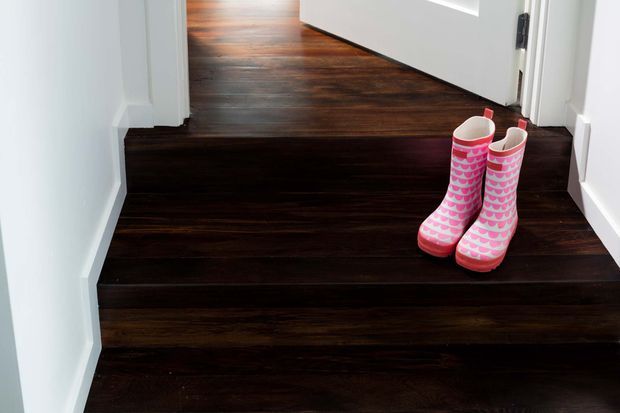 Close-up view of a short interior stairway with dark stained timber treads and adjoining dark timber flooring. The walls, skirting boards, and door frames are white, creating a strong contrast. A pair of bright pink patterned children's gumboots sits on the upper step.