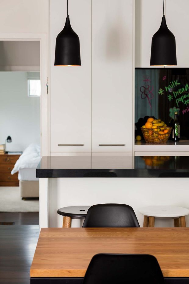 A contemporary kitchen and dining area with white full-height cabinetry, a dark stone island benchtop, black pendant lights, and a warm timber dining table with black chairs. A chalkboard-style splashback or display panel and fruit bowl add visual interest, while an open doorway reveals part of an adjacent bedroom or living space.