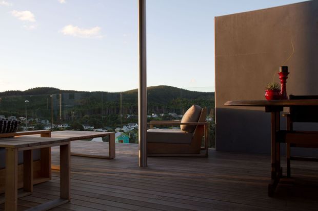 A contemporary outdoor deck or terrace overlooking a wooded, hilly landscape. The space features wide timber decking, a built-in timber dining table and bench, a lounge chair, a slim metal column, and a dark sculptural outdoor dining or console setting against a warm-toned wall. Large open glazing or balustrade panels frame the view.