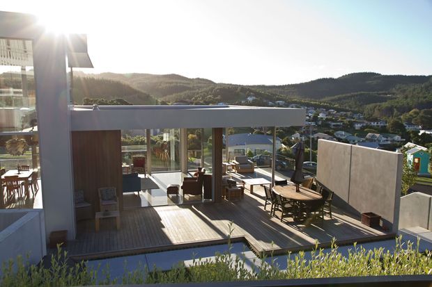 Elevated outdoor terrace and adjoining glass-walled living area with timber decking, outdoor dining furniture, a concrete privacy wall, and expansive views over a hilly suburban landscape. Strong sunlight and extensive glazing create a bright, open connection between interior and exterior spaces.