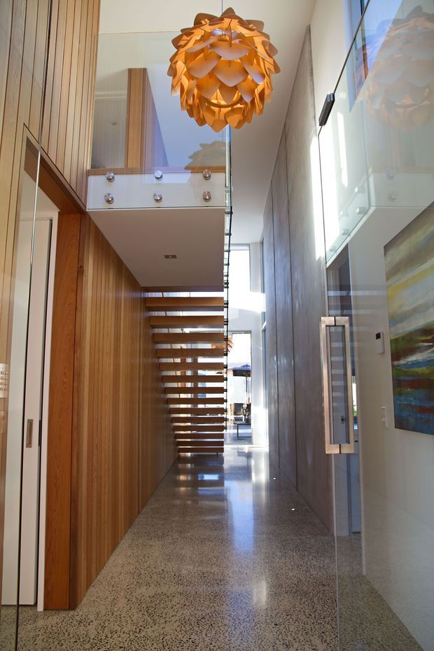 A tall, light-filled residential hallway with a dramatic open floating staircase. The corridor features extensive vertical timber wall panelling and built-in cabinetry, polished speckled concrete or terrazzo flooring, a tall grey feature wall, full-height glazing, and a sculptural warm wood pendant light. A glass balustrade and upper-level landing emphasize the double-height volume.