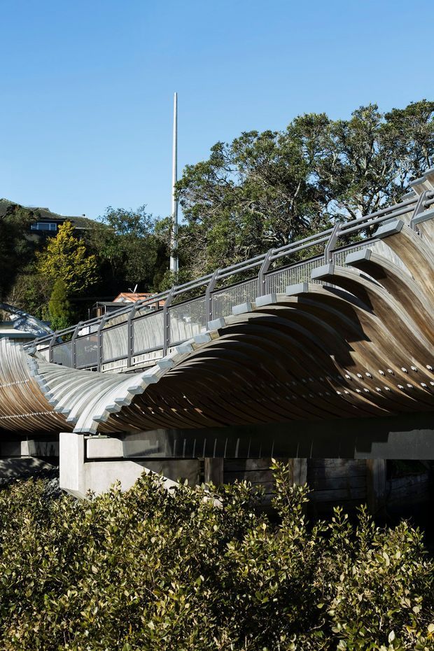 Exterior view of a striking sculptural pedestrian bridge or elevated walkway with repeating curved timber ribs forming a wave-like underside. The structure has grey metal railings, concrete supports, and a landscaped suburban setting with mature trees and nearby houses.