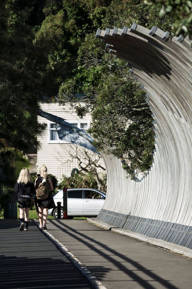 A sunny outdoor pedestrian walkway beside a dramatic curved timber-clad architectural structure, with dense mature greenery, a pale weatherboard building, and pedestrians in the background. The sculptural wall or canopy forms a sweeping tunnel-like curve over the path, creating strong linear shadows and an expressive public-space setting.