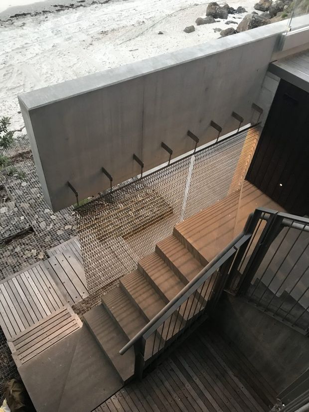 A contemporary exterior stairway and deck viewed from above, with timber decking and stairs, dark metal handrails, and a perforated metal mesh balustrade. A tall smooth concrete privacy wall borders the stair landing, with a dark exterior door at the upper right. Snow-covered rocky terrain is visible beyond the wall.