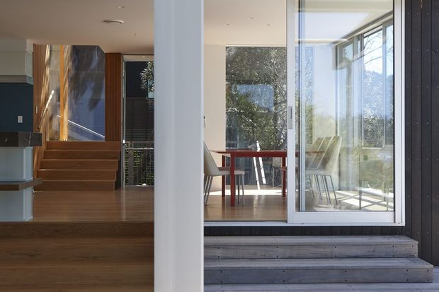A contemporary residential interior-exterior threshold with broad floor-to-ceiling glazing, a timber deck and steps, and a dining area visible inside. The open-plan space includes a red dining table, light chairs, timber flooring and a stairway leading upward beside dark built-in cabinetry. A white structural column and dark vertical exterior cladding frame the view.