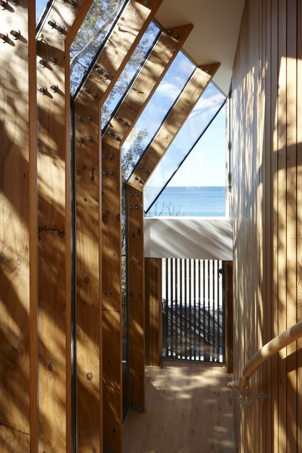 A narrow, sunlit timber stairway or landing with exposed structural wooden members and large angled glazed panels forming a dramatic sloping roof or clerestory. Metal bolts and brackets are visible in the timber framing, while a timber handrail follows the right wall. The view opens toward blue ocean beyond a screened balcony or exterior walkway. Strong natural light creates striped shadows across the timber surfaces and floor.