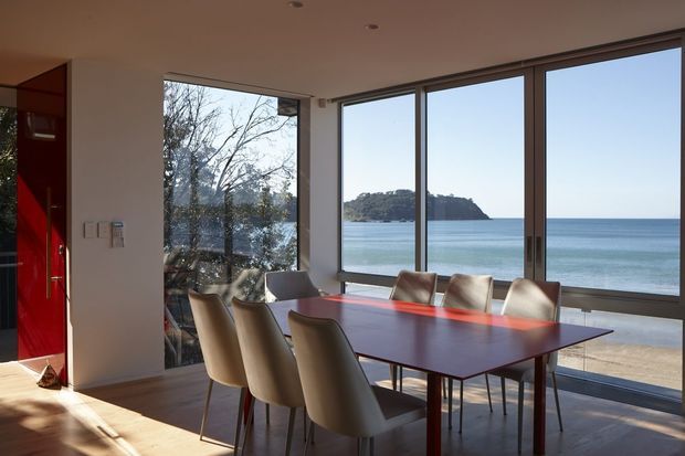 A bright contemporary dining area with a large rectangular timber dining table and upholstered dining chairs, positioned beside expansive floor-to-ceiling windows overlooking a calm coastal bay and sandy beach. The room has a minimalist white interior, warm timber flooring, a bold red entry door, and slim dark window frames.