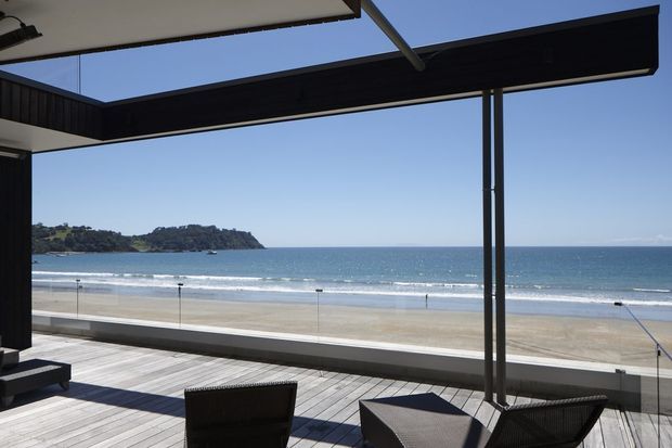 A contemporary beachfront terrace or balcony overlooking a sandy beach, ocean, and headland. The space features dark structural framing, extensive frameless or minimally framed glass balustrading, and pale timber decking with outdoor lounge furniture. A large retractable or louvred overhead structure provides shade while preserving the expansive coastal view.