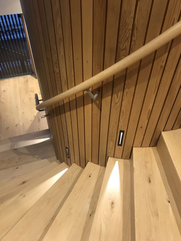 Interior view of a contemporary timber staircase. Broad pale timber treads descend beside a wall clad in narrow vertical natural-wood boards, with a rounded timber handrail on metal brackets and small recessed step lights. Daylight enters from a glazed opening at the stair landing.