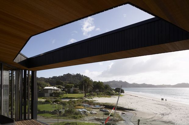View from a covered architectural terrace or balcony toward a sandy beach and bay, framed by a dramatic dark-clad gabled roof structure with a large open skylight-like void. Timber soffits and slender glazed openings define the contemporary coastal building, with landscaped residential surroundings and distant hills beyond the water.