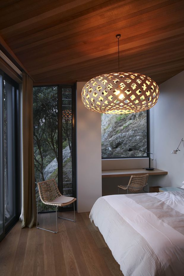 A warm, modern bedroom with a timber-lined ceiling and timber floor, full-height dark-framed glazing, and a large window overlooking a rocky natural landscape. The room contains a bed with white linen, woven lounge chairs, a compact desk or ledge, and a striking woven pendant light. The restrained natural palette and abundant glazing create an intimate, organic atmosphere.