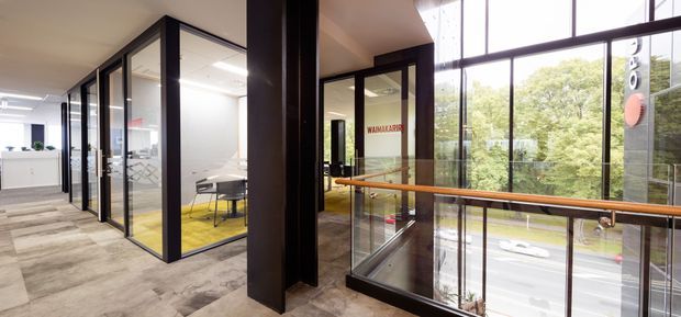 A bright contemporary commercial office interior with an open circulation area, enclosed meeting rooms framed in black metal and glass, and a mezzanine-style balustrade overlooking a street. The space features a polished concrete-look floor, exposed dark structural columns, extensive glazing, and a meeting room with a vivid yellow carpet accent. Trees and vehicles are visible outside through the windows.