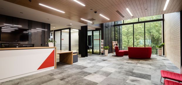A contemporary commercial reception and lounge area with a white reception desk featuring a bold red geometric front panel, dark cabinetry, a timber slatted ceiling, black-framed glazed partitions, and expansive windows overlooking greenery. Grey patterned carpet tiles cover the floor, while red lounge seating and indoor plants provide accents.