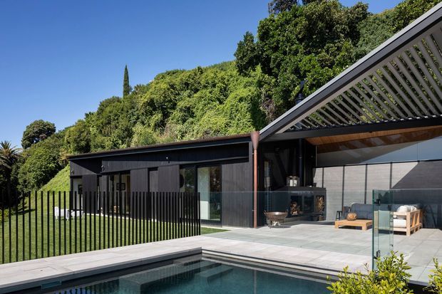 Contemporary single-storey residence with dark vertical cladding, extensive glazing, and a large slatted outdoor canopy over a paved entertaining area. The home opens onto a swimming pool, with landscaped greenery surrounding the property.