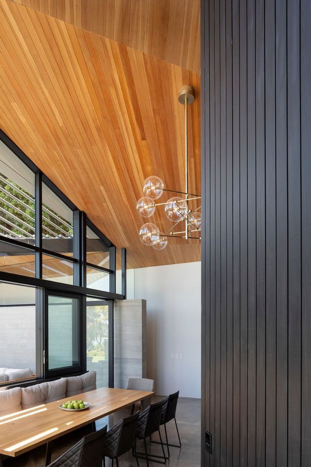 A contemporary dining area viewed beneath a dramatic high, timber-lined ceiling. The room features a long timber dining table with upholstered and black chairs, a sculptural multi-globe pendant light, extensive black-framed glazing, pale walls, and a prominent dark vertical timber feature wall or screen. A glimpse of an adjacent living area is visible through the glazing.