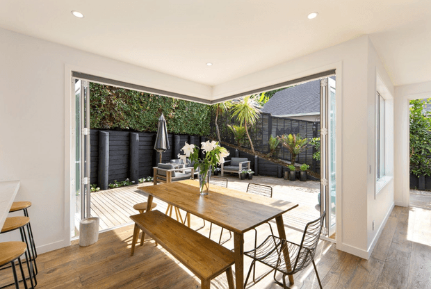 Bright contemporary dining area opening through fully glazed bi-fold doors onto a private timber deck courtyard. The room has pale walls, recessed ceiling lights, warm timber flooring, a large rustic timber dining table with bench seating, and wire-frame chairs. Black fencing, lush planting, and a covered outdoor seating area are visible beyond.
