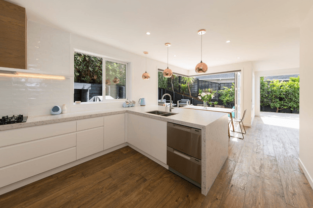 Bright contemporary kitchen with a large L-shaped white island and integrated stainless-steel dishwasher, white gloss tiled splashback, pale stone benchtops, white cabinetry, and warm timber flooring. Three copper pendant lights hang above the island, while large windows and glazed doors open the room toward a leafy outdoor area.