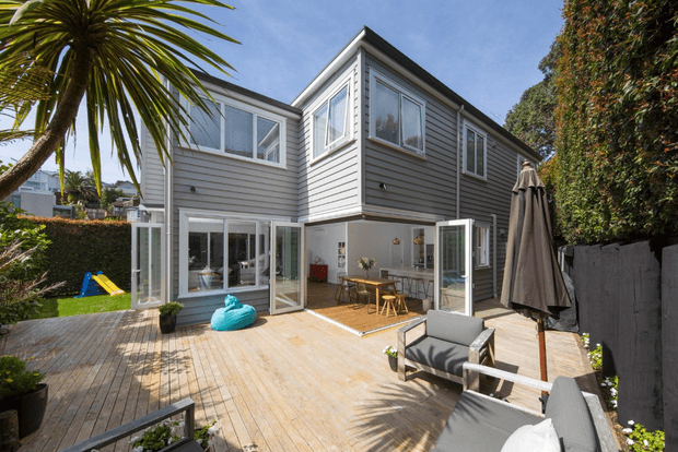 A sunny rear exterior view of a two-storey home with grey horizontal weatherboard cladding, white-framed windows, and extensive glazed openings. The house opens onto a large timber deck with outdoor seating, a dining area, umbrella, plants, and a small children’s slide visible in the garden.
