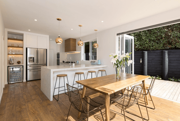 Bright open-plan kitchen and dining area with white cabinetry, stainless-steel appliances, a large stone island with seating, and a timber dining table with wire-frame chairs. Pendant lights hang above the island, while wide glazed doors open to a timber deck and enclosed outdoor area.