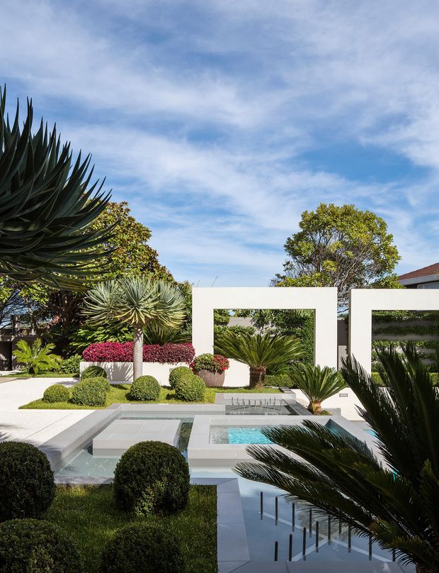 A bright, meticulously landscaped contemporary courtyard garden featuring a geometric reflecting pool and raised square spa, white rendered rectangular garden walls, clipped spherical shrubs, and sculptural palms and succulents. A residential building is partially visible behind the garden.