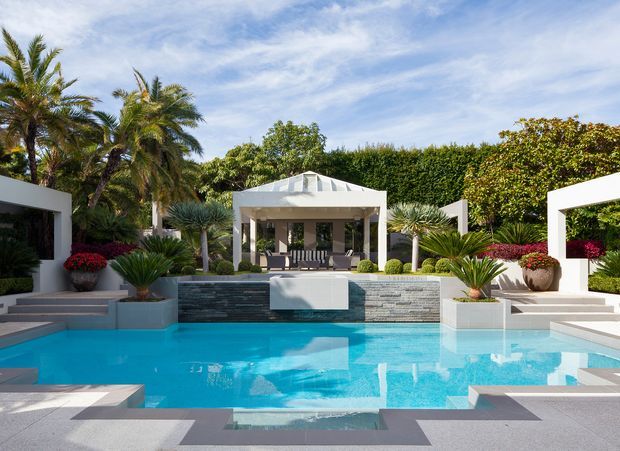 A bright, landscaped contemporary pool courtyard with a large turquoise swimming pool, geometric stepped paving, white rendered pavilion structures, a central covered outdoor lounge, and lush tropical planting including palms and sculptural succulents.