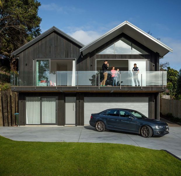 Front exterior view of a contemporary multi-level home with dark vertical timber cladding, two prominent gabled roof forms, large glazed openings, and a glass-fronted upper balcony. A garage and entry level sit beneath the living spaces, with a driveway and car in front.