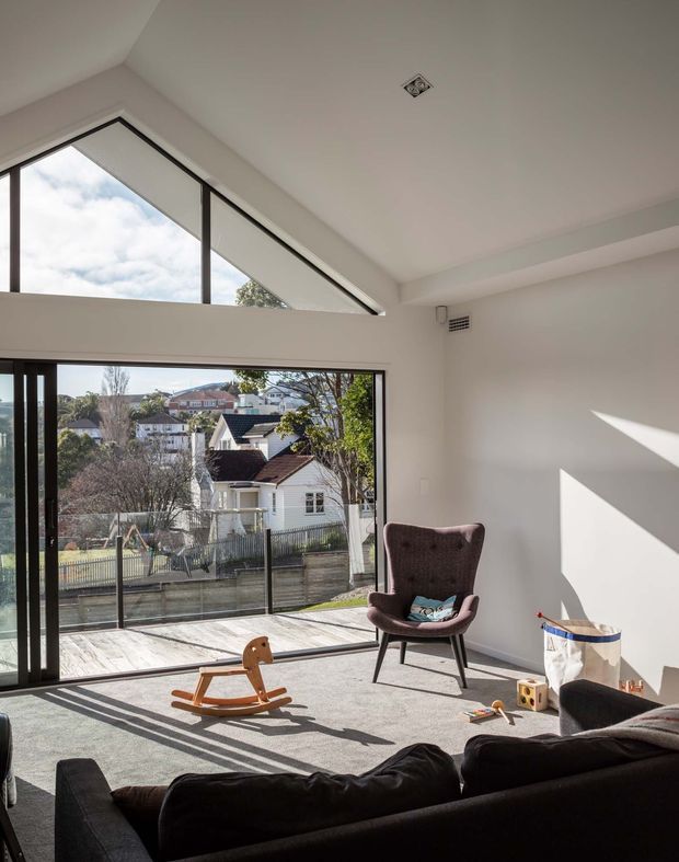 Bright contemporary living room with a high sloping ceiling, large floor-to-ceiling glazing and a triangular clerestory window overlooking neighbouring homes. The room has neutral walls and carpet, a dark sofa, brown upholstered armchair, children's wooden rocking horse and scattered toys, with strong afternoon sunlight casting shadows across the floor.