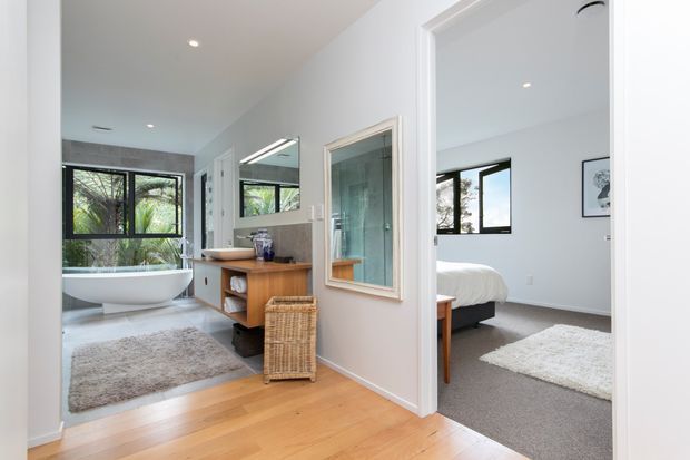 Bright contemporary residential hallway looking toward an open bathroom and bedroom. The bathroom has a freestanding white bathtub, large window, grey rug, timber vanity with integrated basin, and glass shower enclosure. The adjacent bedroom has a white bed, grey carpet, white walls, black-framed window, and a pale rug. Warm timber flooring runs through the hall, with white door and wall trims.
