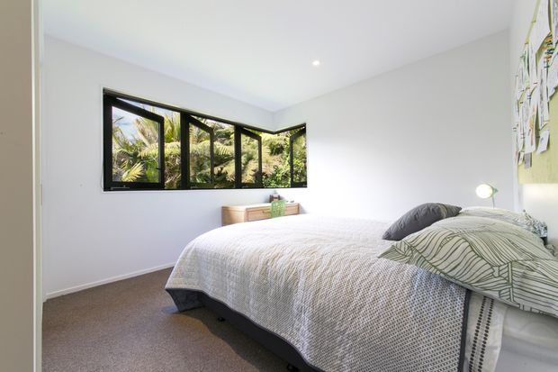 Bright, simply furnished bedroom with a double bed, textured white bedding, patterned pillows, a small timber dresser, and a wall-mounted bedside light. White walls and ceiling create an airy, uncluttered interior, while black-framed corner windows provide views of lush vegetation outside.